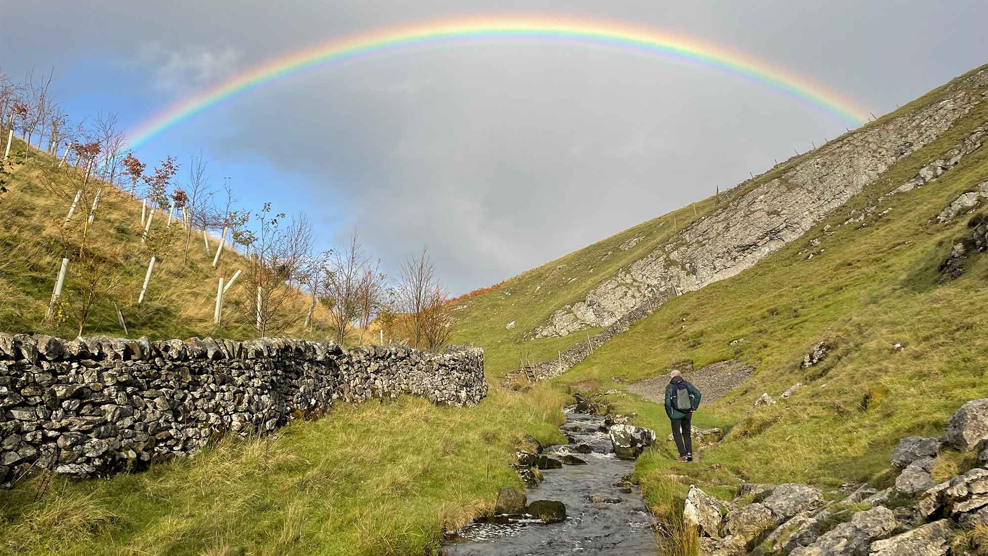 Tollers Gill, Yorkshire. A hiker stood by the stream on a grassy bank  which rises into high into a hill. the other side there is a stone wall. A beautiful rain arches across the sky above